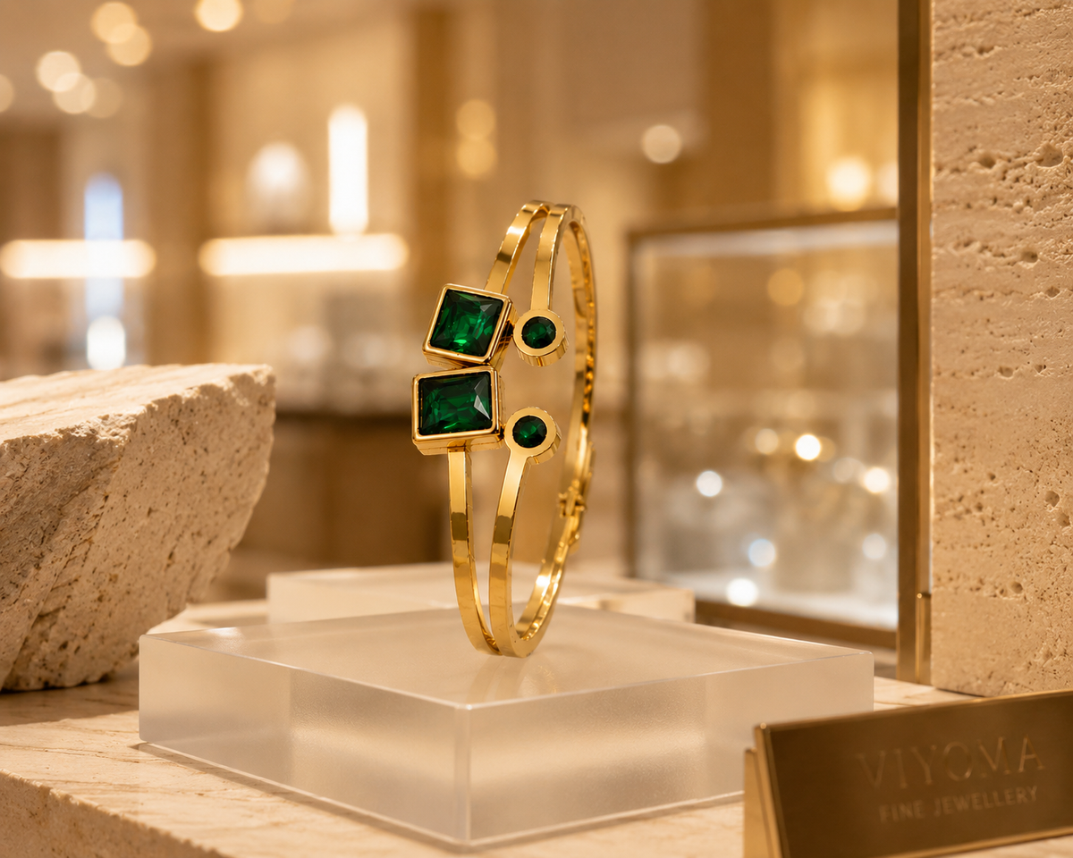 VIYOMA Emerald Royale Cuff featuring emerald-green square and round crystals set on a sleek gold-tone open cuff displayed in a luxury jewelry boutique setting.